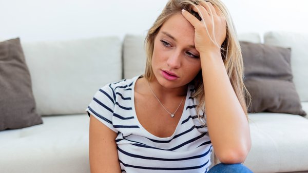Eine junge Frau sitzt erschöpft vor einem Sofa und stützt ihre Stirn in die Hand.
